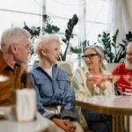 Four seniors sitting together at a retirement home lounge area, engaged in lively conversation while enjoying drinks. They are smiling and laughing, creating a warm and friendly atmosphere that highlights social connections in retirement.