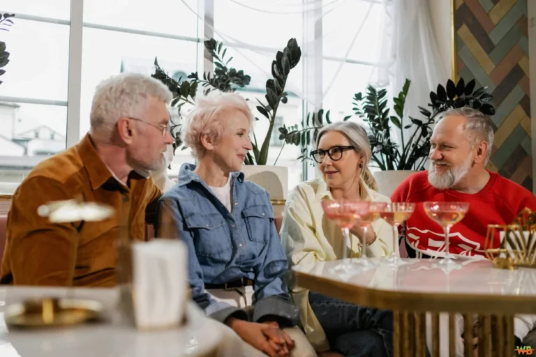 Four seniors sitting together at a retirement home lounge area, engaged in lively conversation while enjoying drinks. They are smiling and laughing, creating a warm and friendly atmosphere that highlights social connections in retirement.