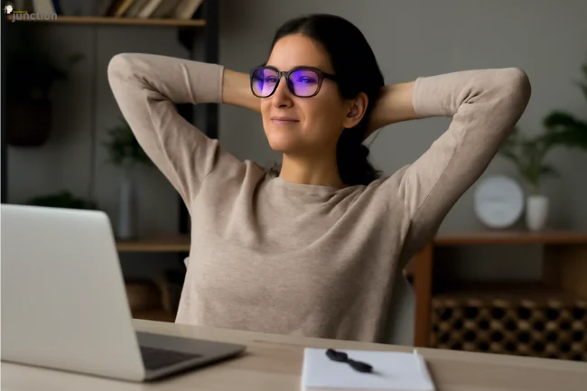 Young woman wearing blue light glasses, taking a break and stretching at her desk to reduce eye strain.
